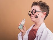 Niño químico en vasos de plástico con manchas de pintura en la cara que huele líquido de la botella sobre fondo beige - foto de stock