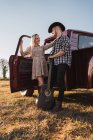 Novio en sombrero de vaquero con guitarra acústica, mientras que de pie con su novia en rojo coche de recogida retro estacionado en la carretera de arena en el campo - foto de stock
