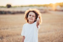 Enfant ethnique joyeux avec des cheveux bouclés montrant comme geste tout en se tenant dans un champ séché en été dans le dos éclairé et cligner des yeux à la caméra — Photo de stock