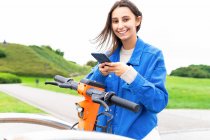 Contenido femenino alquiler de scooter eléctrico estacionado en la ciudad y el teléfono móvil de navegación - foto de stock