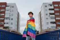 Dal basso di grave omosessuale maschio in piedi con bandiera arcobaleno sul ponte e guardando la fotocamera — Foto stock