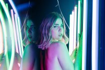 Visão lateral da jovem mulher sonhadora refletindo entre linhas de tubos de néon brilhantes enquanto olha para a câmera — Fotografia de Stock