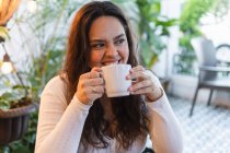 Jovem de cabelos longos latino-americano fêmea desfrutando delicioso café aromático de xícara de cerâmica, enquanto descansa em café acolhedor com plantas verdes — Fotografia de Stock