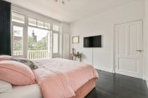Дизайн інтер'єру будинку простора спальня з білими стінами і дерев'яною підлогою, обставлена зручним ліжком з рожевим покривалом і подушками і прикрашена квітами і макетним малюнком в денне світло — стокове фото