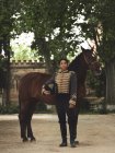 Volle Länge der erwachsenen schwarzen Hündin in elegantem Outfit, die mit braunem Pferd in der Nähe von grünen Bäumen und Burgzaun am Tag im Hof steht — Stockfoto