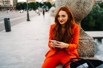 Mujer elegante feliz con el pelo rojo y en traje naranja vibrante sentado mirando a la cámara en el borde de piedra en la ciudad y mensajería en el teléfono celular - foto de stock