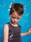 Preteen garçon regardant loin en studio avec des bulles de savon volant sur fond bleu — Photo de stock