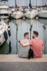 Vista trasera del perro entre el hombre barbudo alegre abrazando la pareja homosexual anónima mientras habla y se sienta en el muelle en el puerto - foto de stock