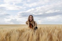 Young female with wavy hair looking at camera bending forward in countryside field under cloudy sky on blurred background — Stock Photo