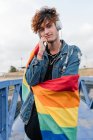 Homosexual elegante hombre con colorida bandera LGBT de pie en el puente y escuchar música en los auriculares mientras mira a la cámara - foto de stock