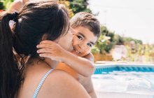 Visão traseira da mãe irreconhecível com filho alegre em pé na piscina perto de árvores verdes no dia ensolarado de verão — Fotografia de Stock