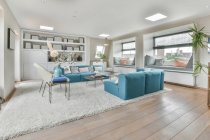 Дизайн интерьера открытой жилой площади с синим диваном и стульями, расположенными рядом с маленьким столом на мягком ковре в современной квартире с белыми стенами и потолком, освещаемым лампами — стоковое фото