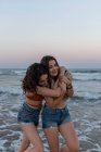 Jóvenes novias alegres abrazándose mientras están de pie en la playa de arena cerca del mar ondeando al atardecer - foto de stock