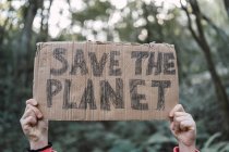 Zugeschnittenes unkenntliches ethnisches Kind zeigt Save The Planet-Titel auf Kartonstück, während es im Wald in die Kamera schaut — Stockfoto