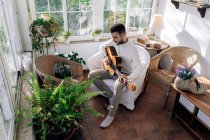 Besinnlicher männlicher Musiker mit Tätowierungen, der klassische Gitarre spielt, während er im Sessel sitzt und gegen das Fenster im Haus blickt — Stockfoto