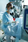 Aufmerksame Ärztin in chirurgischer Uniform und steriler Maske blickt durch das Mikroskop, während sie Auge einer unkenntlichen Patientin im Krankenhaus operiert — Stockfoto