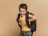 Позитивний крутий дев'ятнадцятикласник з рюкзаком дивиться на камеру на коричневому фоні в студії — стокове фото