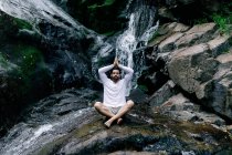 Спокойный мужчина сидит в позе лотоса с молитвенными руками на скале возле водопада и медитирует во время занятий йогой с закрытыми глазами — стоковое фото