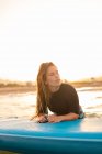 Женщина-серфер лежит на доске SUP и плавает на спокойной воде моря в солнечный день, глядя в сторону во время заката — стоковое фото