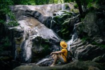 Side view of unrecognizable male hiker sitting on boulder and admiring waterfall in forest — Stock Photo