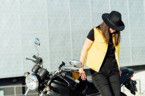 Anonimo motociclista donna in cappello appoggiato sulla moto moderna parcheggiata su strada in città nella giornata di sole — Foto stock