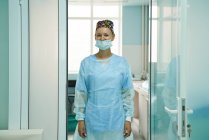 Médico feminino adulto alegre em máscara estéril e boné ornamental olhando para a câmera no hospital — Fotografia de Stock