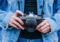 Crop photographe femelle méconnaissable en jean et veste en denim debout avec appareil photo vintage — Photo de stock