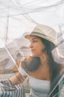 Though transparent curtain of content young female in sunhat chilling in backyard tent on sunny day and looking away — Stock Photo