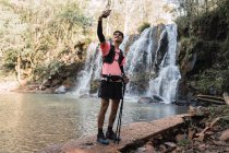 Улыбающийся мужчина-турист делает самоснимок на смартфоне, стоя на фоне водопада и озера в лесу во время похода — стоковое фото