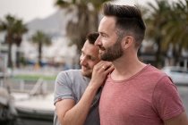 Contenido pareja de hombres homosexuales en camisetas abrazando mientras mira hacia otro lado en el puerto - foto de stock