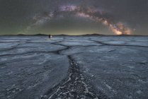 Entfernte Silhouette eines Forschers, der in einer trockenen Salzlagune vor dem Hintergrund des Sternenhimmels mit glühender Milchstraße in der Nacht steht und eine Taschenlampe hält — Stockfoto