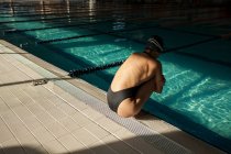 Jeune belle femme à l'intérieur de la piscine intérieure, portant un maillot de bain noir, accroupi sur le bord, vue latérale — Photo de stock