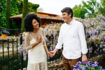 Contenida pareja multirracial cogida de la mano mientras caminan en el jardín con flores de glicina púrpura florecientes y disfrutando el fin de semana juntos - foto de stock