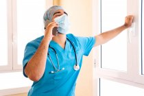 Male medic in uniform and with stethoscope standing in hospital and talking on smartphone while looks up — Stock Photo