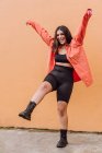 Corpo cheio de alegre millennial feminino na roupa elegante chutando ar contra a parede laranja — Fotografia de Stock