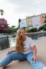 Щаслива стильна жінка з довгим волоссям сидить на бетонній набережній в екзотичному місті влітку і дивиться на камеру — стокове фото