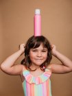 Веселая девочка-подросток в купальнике, стоящая с бутылкой крема от загара на голове на коричневом фоне в студии и смотрящая в камеру — стоковое фото