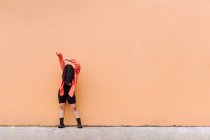 Full body of young anonymous female covering face with long brown hair raising arms while standing against orange wall — Stock Photo