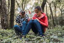 Chica étnica escribiendo en bloc de notas contra hermano mirando a través de binoculares mientras está sentado en la tierra en los bosques de verano - foto de stock