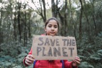 Bambino etnico che mostra il titolo Save The Planet sul pezzo di cartone mentre guarda la fotocamera nella foresta — Foto stock