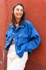 Позитивна жінка в стильному вбранні, яка дивиться, спираючись на барвистий фон стіни будівлі в сонячний день на міській вулиці — стокове фото