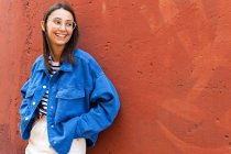 Positive Frau in stilvollem Outfit schaut weg, angelehnt an bunten Hintergrund der Hauswand an einem sonnigen Tag in der City Street — Stockfoto