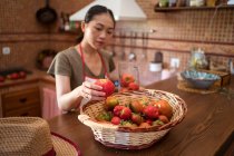 Enfocado ama de casa étnica que pesa tomates frescos en jarra de vidrio en la balanza de la cocina mientras cocina alimentos en casa - foto de stock