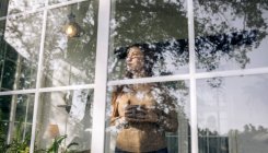 Durch den Fensterblick einer nachdenklichen jungen Frau mit einem Becher Heißgetränk, die gegen das Fenster im Haus blickt — Stockfoto
