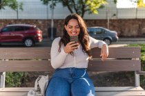 Jovem e alegre estudante latino-americana navegando no celular enquanto descansa no banco de madeira na rua da cidade no dia de verão — Fotografia de Stock