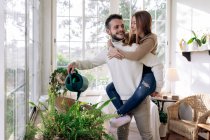 Smiling woman riding piggyback on boyfriend watering plant while looking at each other against windows at home — Stock Photo