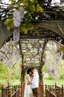 Vista lateral de una suave pareja multirracial abrazándose mientras está de pie con los ojos cerrados bajo el arco con flores de glicina violeta florecientes en el jardín - foto de stock