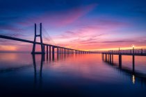 Silueta de Vasco da Gama Puente y largo muelle situado en el tranquilo río Tajo contra el cielo nublado al atardecer en la noche en Lisboa, Portugal - foto de stock