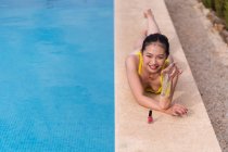 De cima Asiática fêmea em biquíni amarelo deitado perto da piscina e banhos de sol enquanto desfruta de férias de verão no resort — Fotografia de Stock