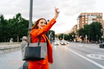 Позитивная рыжая женщина в оранжевом костюме вызывает такси с поднятой рукой, стоя на обочине дороги в городе и отводя взгляд — стоковое фото